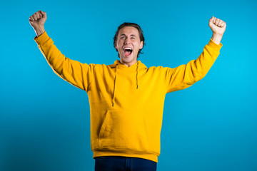 Fototapeta premium Yes winner gesture. Happy european man. Handsome guy with stylish hairdo surprised to camera over blue background