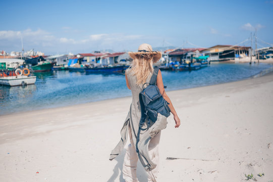 Traveler blonde backpacker woman in straw hat back view walks along the coast at the pier fishing boats. Travel adventure in China.Tropical island Asia tourist. Summer holiday vacation journey concept