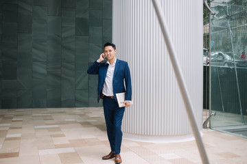 Positive young modern Asian businessman with laptop standing and speaking over smartphone