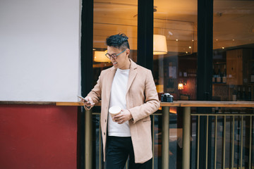 Glad ethnic casual guy in coat enjoying in takeaway coffee while surfing on smartphone at street