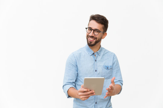Happy joyful guy with tablet getting good news, looking at camera, smiling, gesturing. Handsome young man in casual shirt and glasses standing isolated over white background. Good news concept