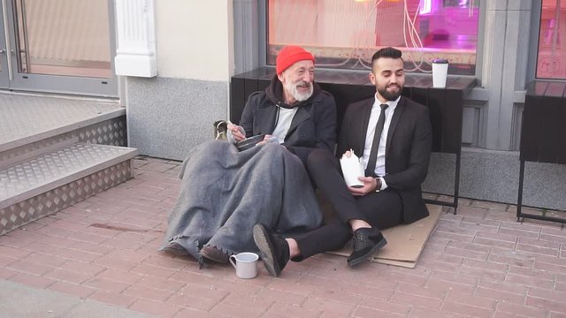 Smiling rich and poor men together sitting on street and eat while speaking. Happy despite social inequality