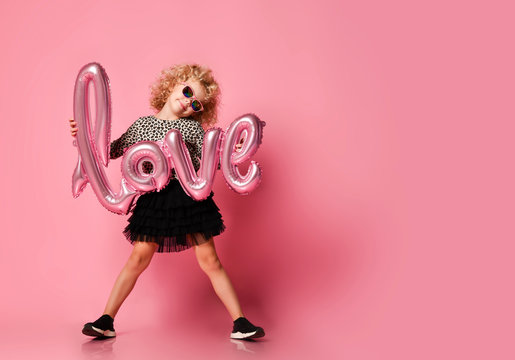 Smiling kid girl in leopard print shirt and fatin skirt is posing with love word air ballon on pink with copy space