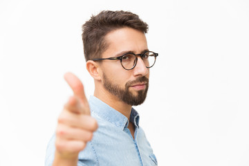 Closeup of serious guy pointing finger at camera. Handsome young man in casual shirt and glasses standing isolated over white background. Choosing you concept
