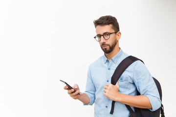 Serious cellphone user with backpack surfing internet, looking at camera. Handsome young man in casual shirt and glasses standing isolated over white background. Wireless technology concept