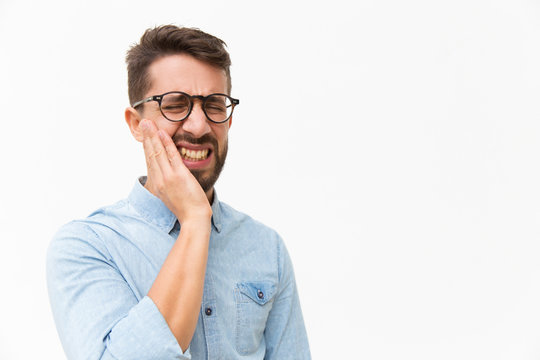 Upset Unhappy Guy Holding Cheek With Pain Face. Handsome Young Man In Casual Shirt And Glasses Standing Isolated Over White Background. Suffering From Tooth Ache Concept