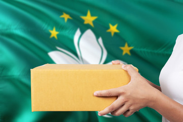 Macao delivery service. International shipment theme. Woman courier hand holding brown box isolated on national flag background.