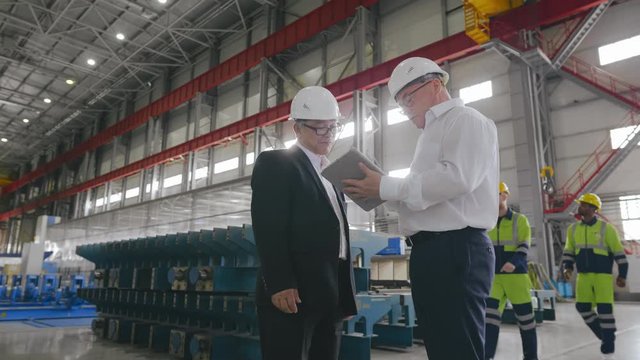 Businessman and engineer talk looking checking blueprints. Two business people or contractors discussing project with documents standing in large workshop of modern factory