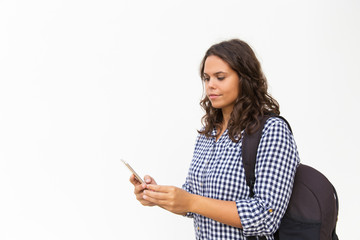 Fototapeta premium Focused female tourist with backpack and cellphone using location app. Young woman in casual checked shirt standing isolated over white background. Tourist with smartphone concept