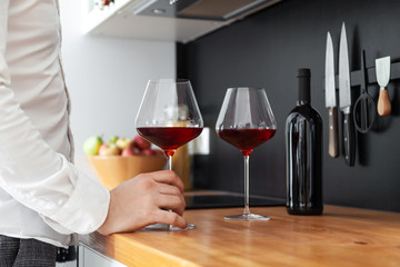 man in white shirt holds in his hand burgundy glass with red wine, bottle stands on wooden tabletop of Scandi kitchen against a black wall, knives. Concept romantic dinner at home, date, party