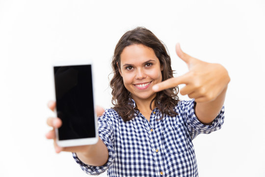 Happy positive customer pointing finger at blank phone screen. Young woman in casual checked shirt standing isolated over white background. Advertising or mobile app concept