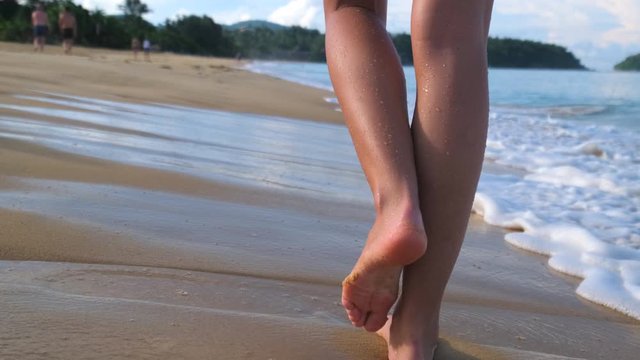 Beach travel - woman walking barefoot on beach coastline leaving footprints in sand. Closeup of tanned female feet legs on ocean sea. wave footsteps tourism relax tropical happy holiday. slow-mo 4 K