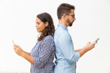 Couple standing back-to-back and using smartphones. Young woman in casual and man in glasses posing...