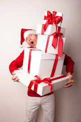 Man wearing Santa hat holding massive pile of Christmas presents looking at the camera with a surprised expression