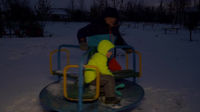 Father and son ride on carousel in park. kid loves to play with his father on holiday. elderly father in bright jacket with gray beard and glasses is pleased to play with baby. Dark winter evening