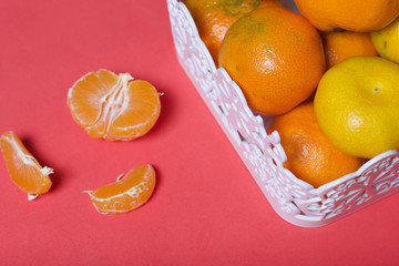 Tangerines in a wicker basket. Next to the basket is peeled mandarin. Visible slices of mandarin and skin from it.  Against the background of coral color.