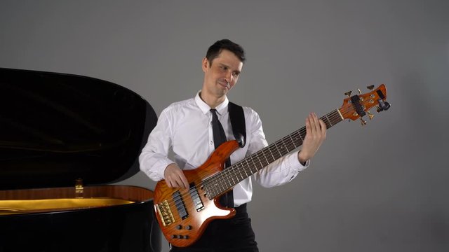 Male musician plays bass guitar on grey background next to piano