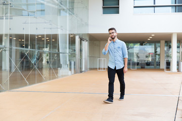 Fototapeta na wymiar Positive guy speaking on cellphone while going through office lobby. Handsome young man in casual shirt and glasses walking indoors with glass wall in background. Phone talk concept