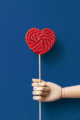 Wooden hand holding a large red and white striped lollipop on blue. The concept of Valentine's Day, love and friendship. Minimal classic flat lay composition layout. Natural material and light