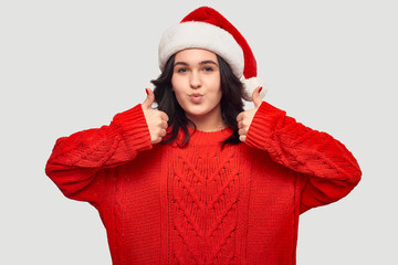 Pretty brunette girl in a red sweater and Santa hat holding thumbs up isolated over grey background