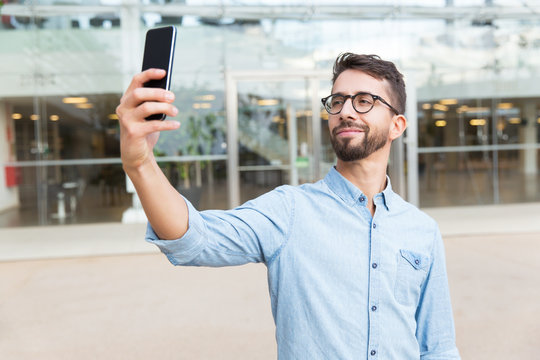 Positive smiling guy in eyewear with mobile phone taking selfie. Handsome young man in casual shirt and glasses standing at outdoor glass wall. Male selfie, leisure, fun concept
