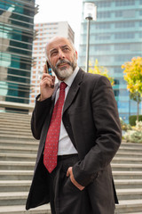 Content businessman talking by smartphone and looking away. Cheerful mature businessman standing with hand in pocket and talking by cell phone on street. Communication concept