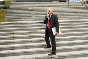 Mature businessman with cell phone and laptop on street. Full length view of businessman holding laptop computer and talking by smartphone while standing on street. Technology concept