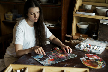 Fototapeta premium Beautiful long-haired woman interferes with a spatula with a palette of oil paints to create a picture. Handwork. Ceramic Making. Wall panel