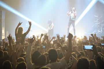 Concert shot, a huge crowd is standing in front of a lit stage clapping a band performance.