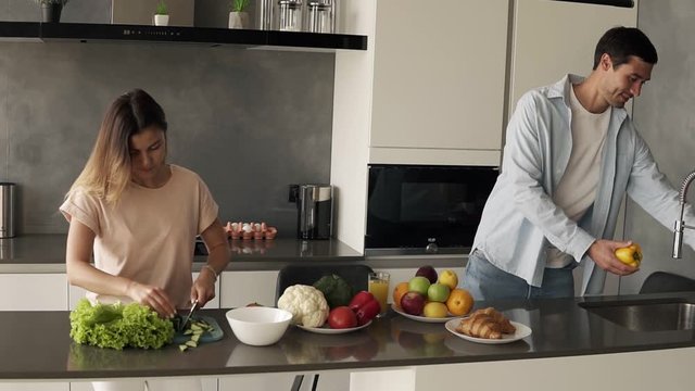 Cheerful young family couple cutting fresh vegetable salad, having fun cooking together im modern loft interior kitchen, happy husband helping prepare healthy meal - washing yellow peper in sink