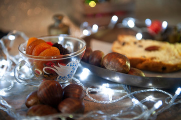 Antique, transparent cup, dried fruits. Focused damasco. Brazilian Christmas table. Portuguese chestnut, panettone, defocused. Selective focus. White Christmas Lights. Seen up close. Horizontal