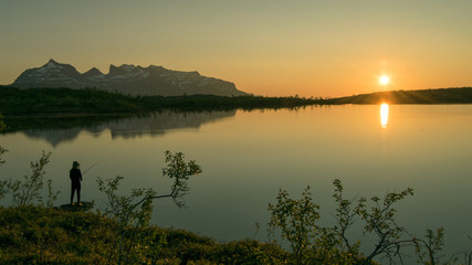 Silhouette of a fishing person in front of a mountain lake, with the midnight sun reflecting in the...