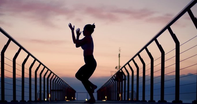Silhouette of girl doing Garudasana pose practicing yoga at amazing early morning sunrise, strong athletic female standing on leg for body stretching on sea beach pier for recreation exercise