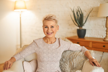 People, lifestyle and mature age concept. Horizontal image of good looking confident female with short dyed hair relaxing at home, sitting comfortably in armchair, smiling broadly at camera
