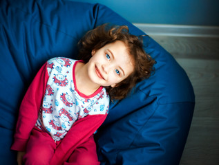 6 years girl child in pink pajamas lay on blue ottoman and looks up with smile. day of sleep. Happiness, leisure and lifestyle in childhood. Natural daylight