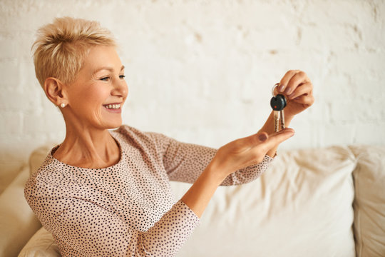 Close up image of happy beautiful European 50 year old woman rejoicing at New Year’ s gift, holding keys to automobile, being excited. Joyful middle aged lady sitting on couch with house key