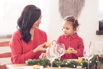 Cheerful mother and her daughter laughing, having fun with Christmas tree and star shaped cookies at the kitchen table. They are in red clothes wearing reindeer antlers and Santa hat headbands.