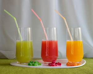 Transparent smooth glasses with colorful juice with stuck tubes are on table. Fruits from which juice is made lie next to pieces. Healthy diet. 