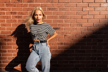 Beautiful blonde girl with loose hair and red lips dressed in grey t-shirt and jeans stands in the sun, holds her hands on her waist, isolated over red brick wall