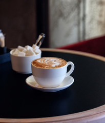 Cappuccino cup of coffee with sugar on the table in restaurant side view and a lot of copy space. Coffee cup latte and saucer. Espresso in cafe, dark background.