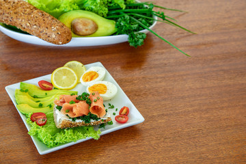 A sandwich with salmon, cream cheese, homemade bread and parsley on a white plate with avocado, salad, lemon and cherry tomato, wooden table
