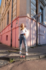 Street style portrait of the beautiful blonde teen girl in white top and blue jeans stands near the corner of the pink old house