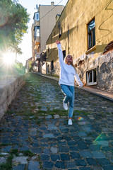 Obraz na płótnie Canvas Street style fashion portrait of the beautiful blonde girl in white hoodie walks along the paving stones road of the old european city, sunset on the background