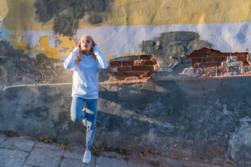 Fototapeta premium Attractive blonde girl in white hoodie leaned on the old grey wall and straightens her hair