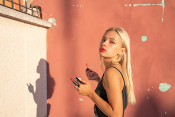 Waist up portrait of attractive young girl in black top that aints her lips with red lipstick, pink wall on the background