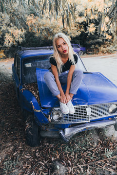 Street style fashion portrait of the blonde girl in black top and blue jeans that sits on the brocken blue old retro car and looking at the camera