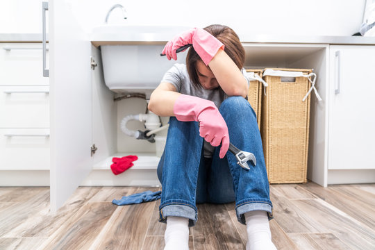 Sad woman sitting near leaking sink in laundry room holding adjustable wrench
