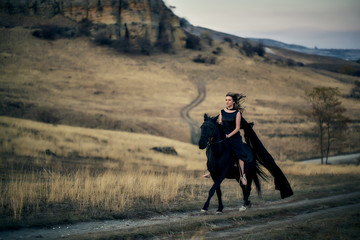 cheerful young beautiful girl in a light flying black dress riding on a black horse gallops on a field against the backdrop of mountains and sunset