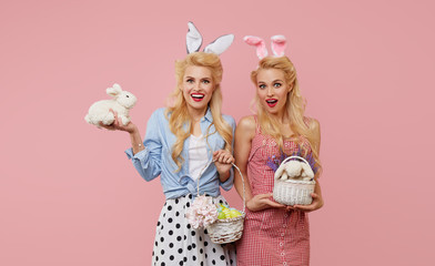 Young pin up women tweens in retro clothes with bunny ears holding a basket full of Easter eggs and string bag with vegetables on pink background. Eco-friendly packaging and zero waste