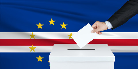 Election in Cape Verde. The hand of man putting his vote in the ballot box. Waved Cape Verde flag on background.